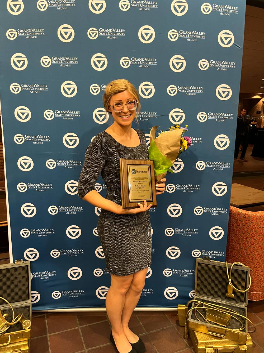 Alum standing in front of the GV Alumni Relations backdrop with an award and flowers.
